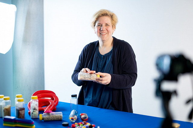 Eine Fortbildnerin steht mit freundlichem Lachen an einem blauen Tisch. Sie probiert an einem Stabmagneten, ob ein Gegenstand magnetisch ist. Im Hintergrund steht eine große Leuchte und eine Kamera. Sie hält einen Webcast zu naturwissenschaftlicher Bildung für pädagogische Fachkräfte aus der Krippe.