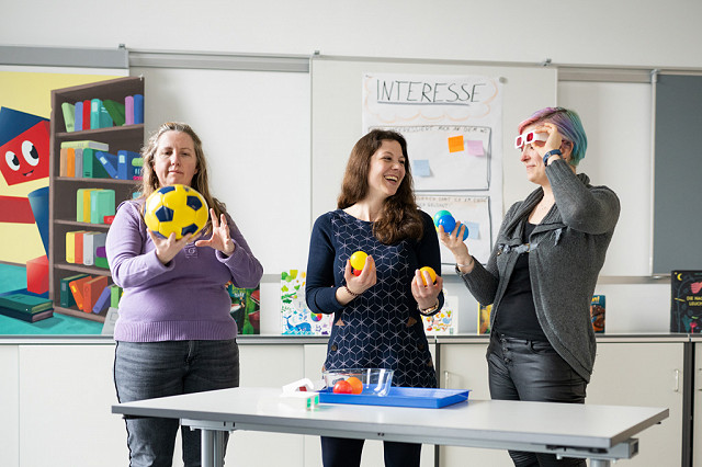 An einem Tisch stehen zwei Erzieherinnen und eine Fortbildnerin während einer Forscherstation-Fortbildung zu früher naturwissenschaftlicher Bildung. Sie schauen sich Bälle in unterschiedlichen Farben an und unterhalten sich dazu.